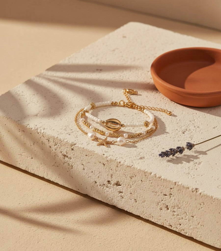 Set de tres tobilleras Clarvyna doradas con concha, estrella de mar y perlas sobre base de piedra rústica.