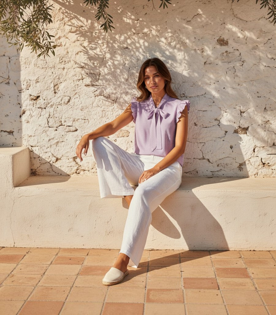 Blusa Clarvyna lila de lunares con volantes y lazo al cuello, combinada con pantalones blancos en un entorno exterior.