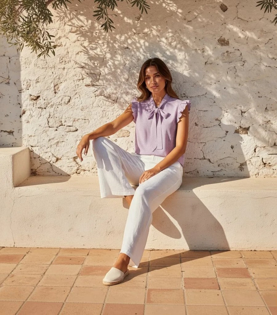 Modelo vistiendo blusa Clarvyna lila de lunares con volantes y lazo al cuello, combinada con pantalones blancos