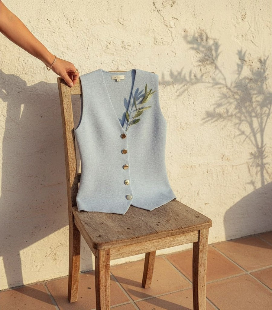 Chaleco de punto Clarvyna azul claro sin mangas con botones frontales sobre una silla de madera al sol