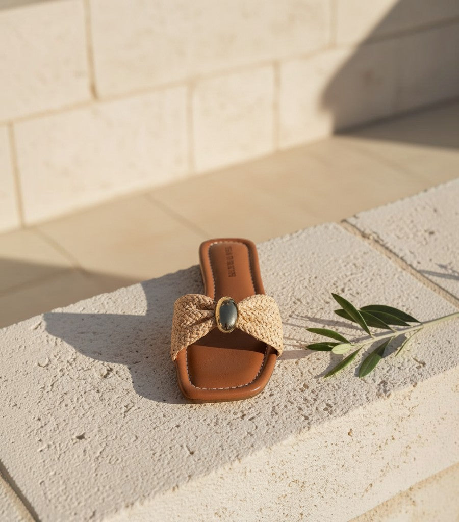 Sandalias planas Clarvyna con tira trenzada color beige, detalle central metálico y plantilla marrón sobre piedra.