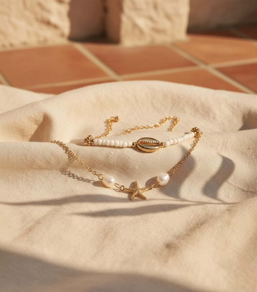 Set de tobilleras Clarvyna doradas con motivos de estrella de mar, concha y perlas sobre tela beige al sol